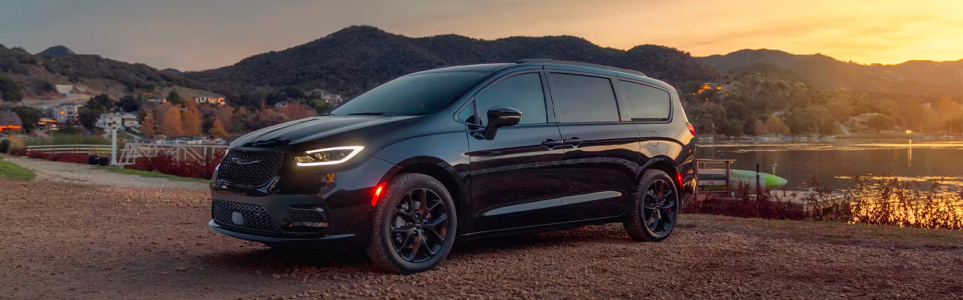 A black Chrysler Pacifica minivan is parked on a dirt road near a lake at sunset