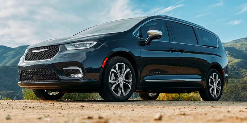 A black Chrysler Pacifica minivan is parked on a dirt road with mountains in the background