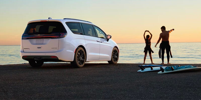 A white minivan is parked on a beach, facing away from a father and child with surfboards