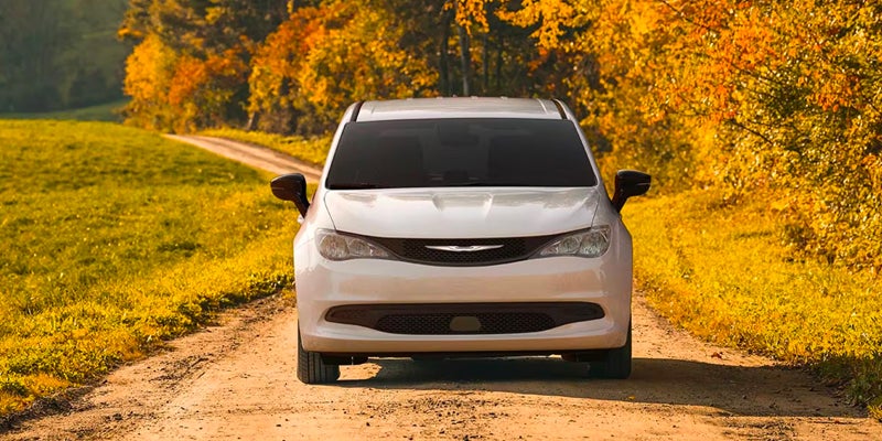 Front view of a white Chrysler minivan driving on a dirt road in autumn
