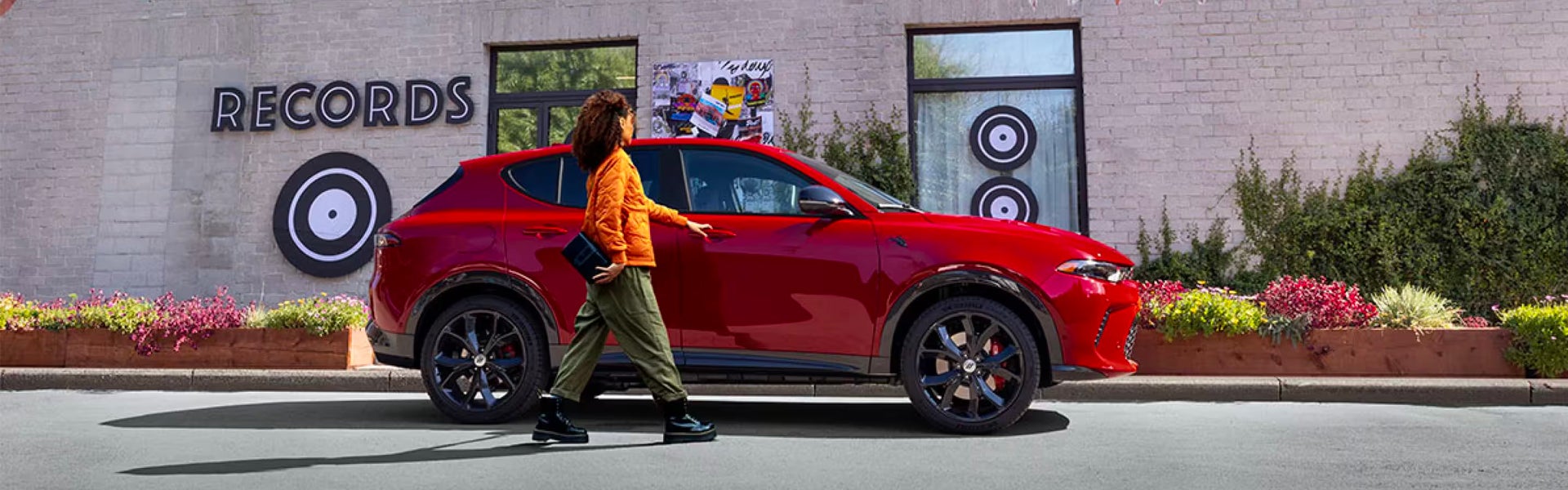 A woman in an orange jacket steps out of a red Alfa Romeo Tonale SUV parked outside a building