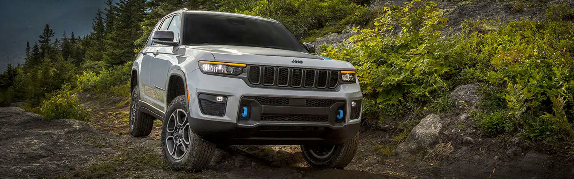 A light gray Jeep Grand Cherokee 4xe SUV with blue tow hooks driving off-road