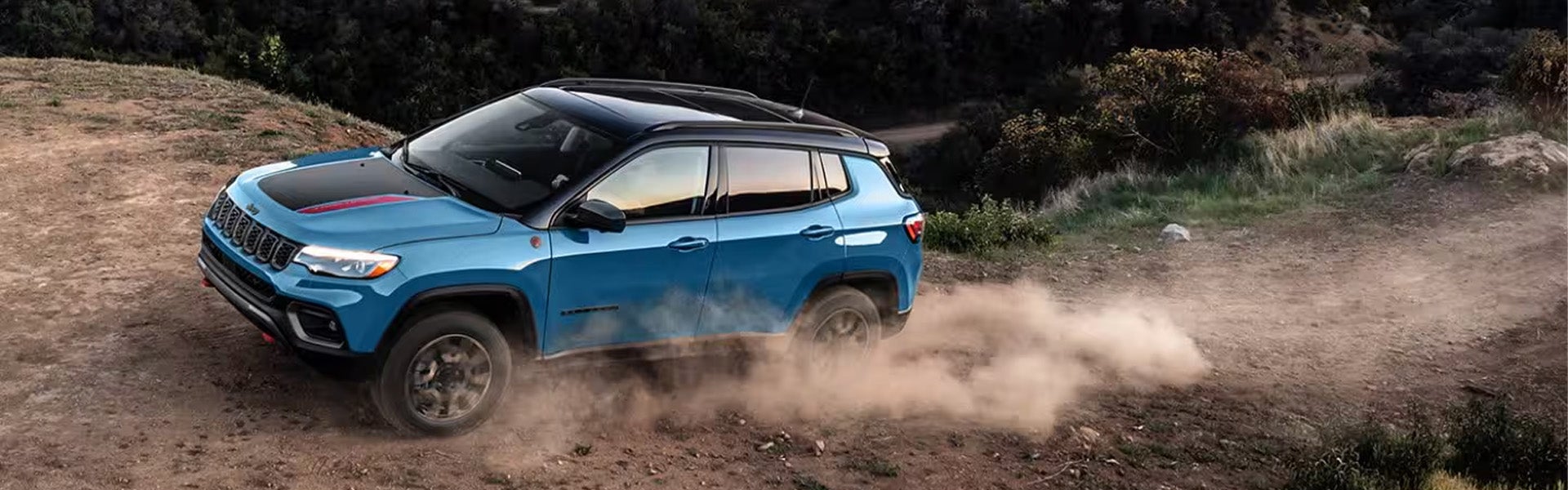 A blue Jeep Compass Trailhawk SUV drives on a dirt road, kicking up dust