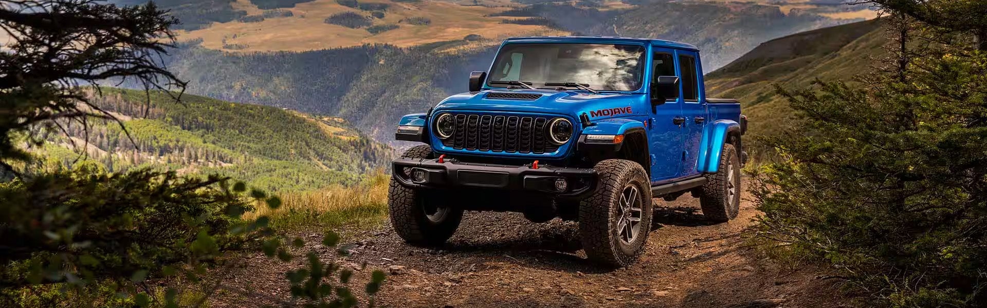 A blue Jeep Gladiator Mojave pickup truck driving on a dirt trail in a mountainous landscape.