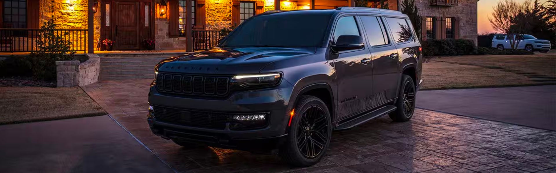 A dark-colored, large SUV parked on a paved driveway in front of a luxury home at dusk