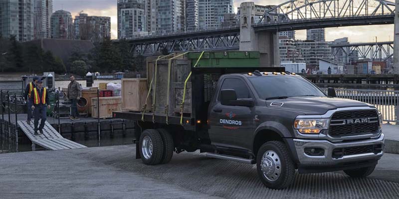 A Ram Heavy Duty flatbed dually truck with cargo, parked near water with a city skyline in the background