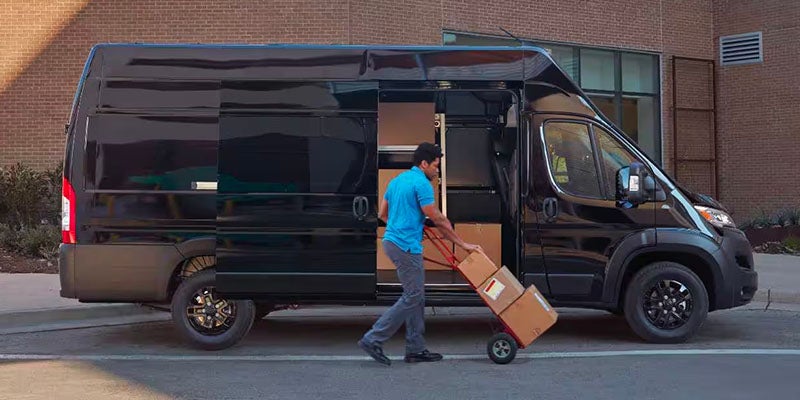 A person in a blue shirt loads boxes into a black Ram ProMaster van